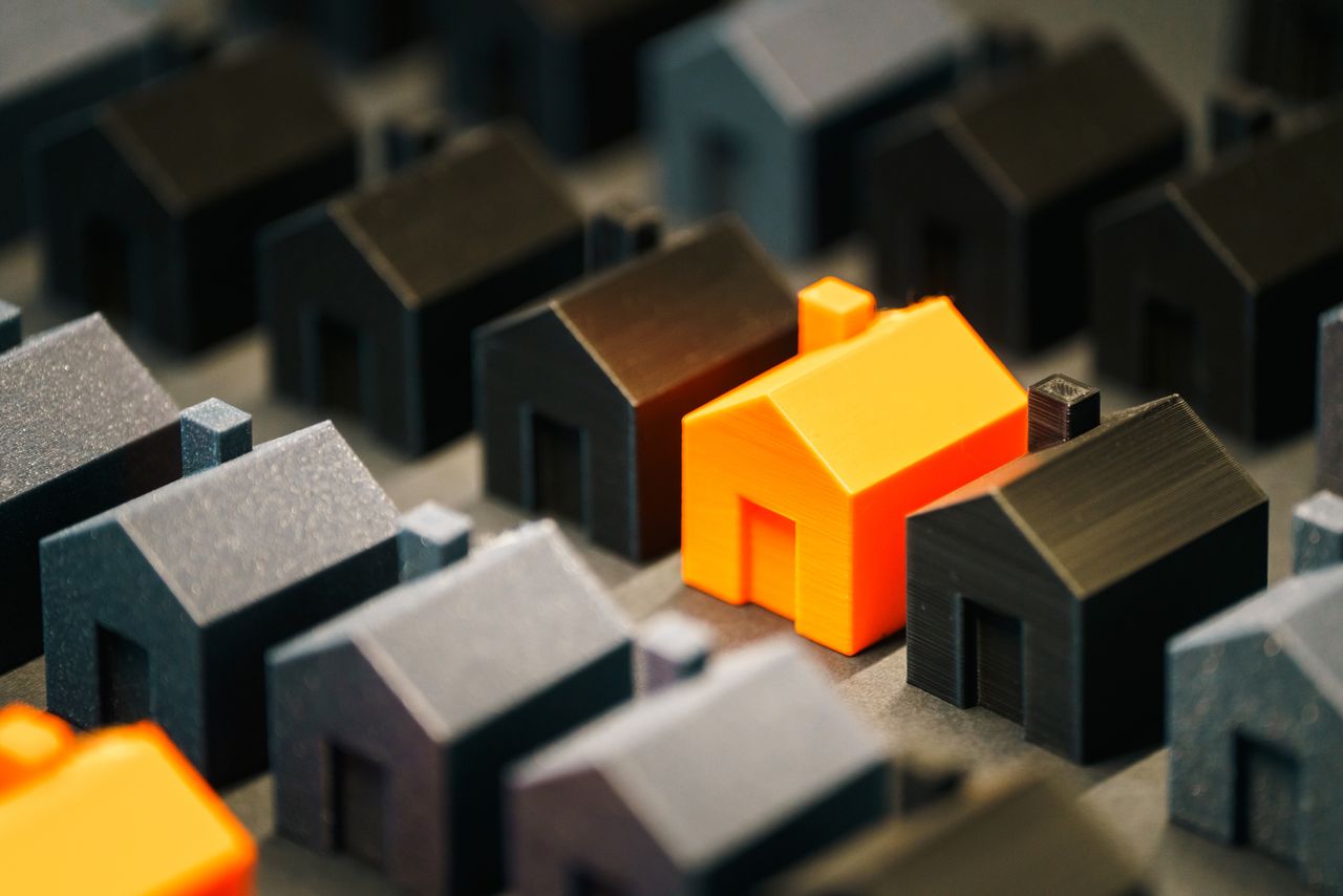 story-img Striking orange model house stands out among black miniature houses, symbolizing real estate choices.